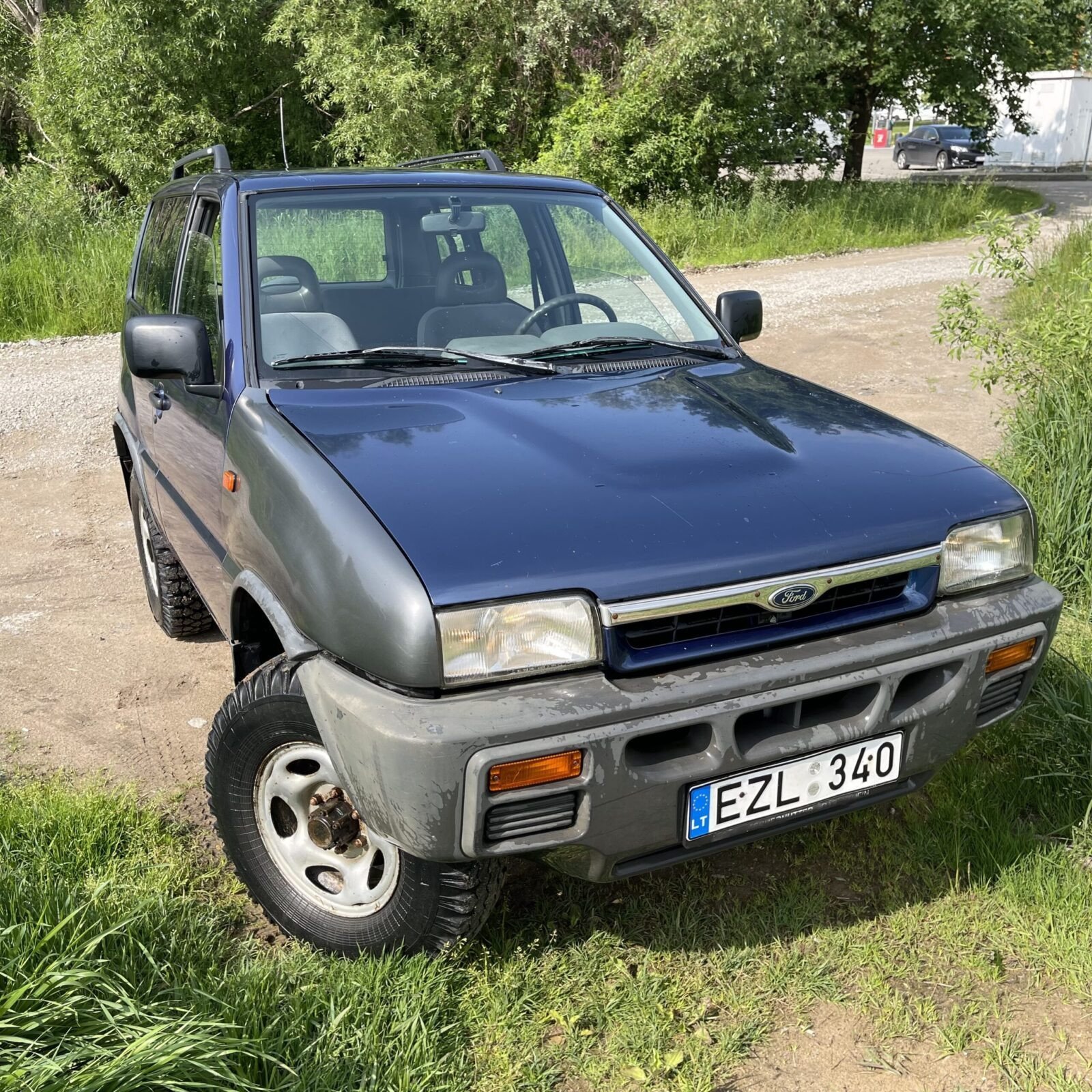 Tamsiai mėlynas Ford Maverick visureigis, pastatytas ant žolės šalia žvyrkelio, iliustruoja automobilių supirkimo paslaugas Klaipėdoje, kurias teikia AutoDylas.