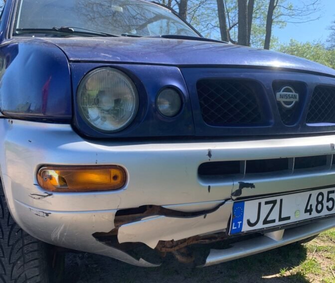 Mėlynas Nissan visureigis su sudaužytu priekiniu bamperiu po virtimo ant šono.