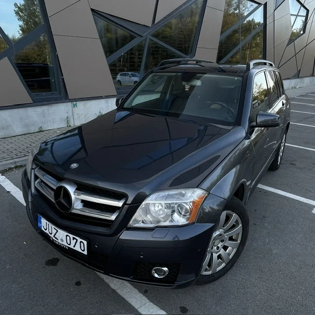 Tamsiai pilkas Mercedes-Benz GLK klasės visureigis, stovintis automobilių stovėjimo aikštelėje prie modernaus pastato.